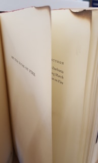 An open book with visible text on two of its pages, showing titles and names. The edges of the pages are slightly browned, possibly indicating age or exposure to heat.