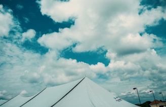 A large white tent is set up under a vibrant blue sky filled with fluffy white clouds. Multiple poles support the tent, and some are visible in the background along with parts of a rooftop.