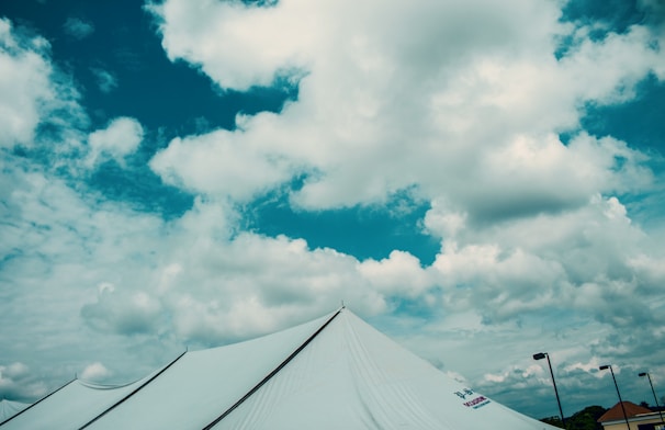 A large white tent is set up under a vibrant blue sky filled with fluffy white clouds. Multiple poles support the tent, and some are visible in the background along with parts of a rooftop.