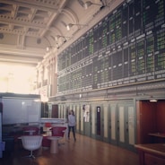 An interior space with a high ceiling featuring a large electronic board displaying various financial data and numbers. There is modern seating with red and white chairs, and a person is walking through the room.