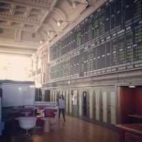An interior space with a high ceiling featuring a large electronic board displaying various financial data and numbers. There is modern seating with red and white chairs, and a person is walking through the room.