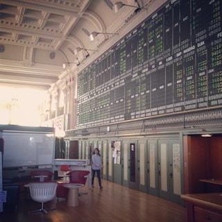 An interior space with a high ceiling featuring a large electronic board displaying various financial data and numbers. There is modern seating with red and white chairs, and a person is walking through the room.