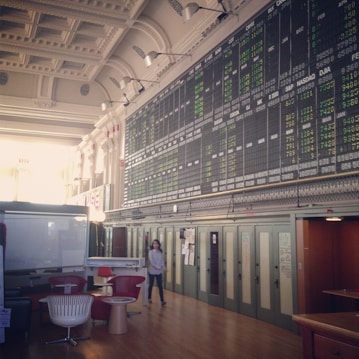 An interior space with a high ceiling featuring a large electronic board displaying various financial data and numbers. There is modern seating with red and white chairs, and a person is walking through the room.
