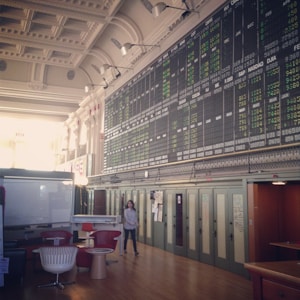 An interior space with a high ceiling featuring a large electronic board displaying various financial data and numbers. There is modern seating with red and white chairs, and a person is walking through the room.