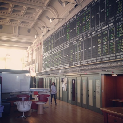 An interior space with a high ceiling featuring a large electronic board displaying various financial data and numbers. There is modern seating with red and white chairs, and a person is walking through the room.