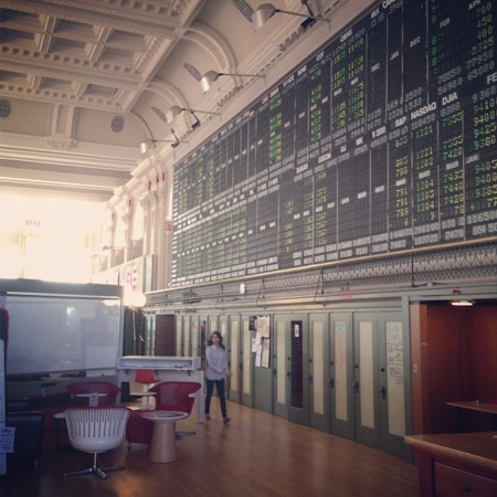 An interior space with a high ceiling featuring a large electronic board displaying various financial data and numbers. There is modern seating with red and white chairs, and a person is walking through the room.