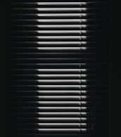 Vertical window blinds are partially opened, casting dramatic shadows and light patterns with strong contrast.
