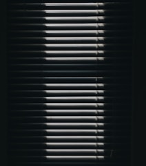 Vertical window blinds are partially opened, casting dramatic shadows and light patterns with strong contrast.