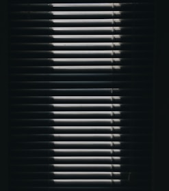 Vertical window blinds are partially opened, casting dramatic shadows and light patterns with strong contrast.
