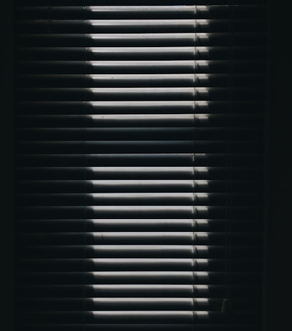 Vertical window blinds are partially opened, casting dramatic shadows and light patterns with strong contrast.