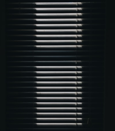 Vertical window blinds are partially opened, casting dramatic shadows and light patterns with strong contrast.
