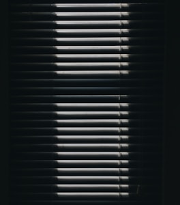Vertical window blinds are partially opened, casting dramatic shadows and light patterns with strong contrast.