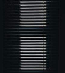 Vertical window blinds are partially opened, casting dramatic shadows and light patterns with strong contrast.