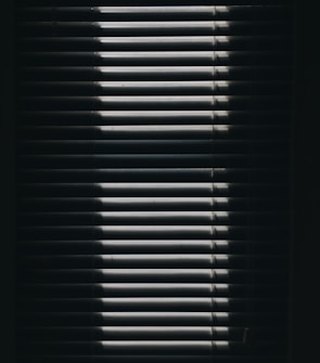 Vertical window blinds are partially opened, casting dramatic shadows and light patterns with strong contrast.