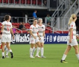 A group of women soccer players in white and gray uniforms are standing on a soccer field. They are wearing matching athletic gear and some are smiling and interacting with each other. The background shows red stadium seats and a few spectators.