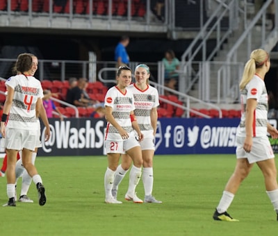A group of women soccer players in white and gray uniforms are standing on a soccer field. They are wearing matching athletic gear and some are smiling and interacting with each other. The background shows red stadium seats and a few spectators.