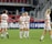 A group of women soccer players in white and gray uniforms are standing on a soccer field. They are wearing matching athletic gear and some are smiling and interacting with each other. The background shows red stadium seats and a few spectators.