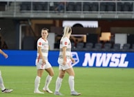 Soccer players in team uniforms stand on a grass field with a large digital advertisement screen displaying the acronym 'NWSL' in the background. The setting appears to be a stadium with rows of empty seats visible above the screen.