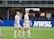 Soccer players in team uniforms stand on a grass field with a large digital advertisement screen displaying the acronym 'NWSL' in the background. The setting appears to be a stadium with rows of empty seats visible above the screen.