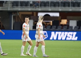 Soccer players in team uniforms stand on a grass field with a large digital advertisement screen displaying the acronym 'NWSL' in the background. The setting appears to be a stadium with rows of empty seats visible above the screen.