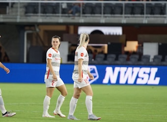 Soccer players in team uniforms stand on a grass field with a large digital advertisement screen displaying the acronym 'NWSL' in the background. The setting appears to be a stadium with rows of empty seats visible above the screen.