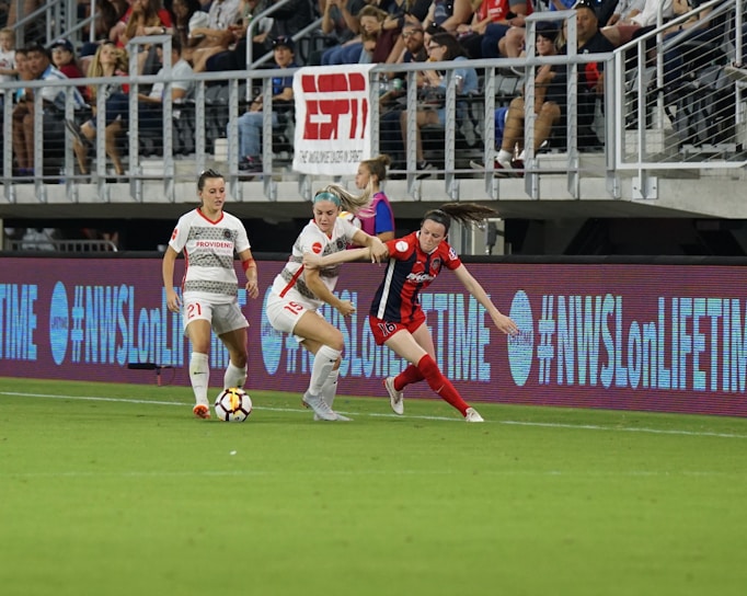 A competitive soccer match is underway with players vying for the ball near the sideline. The players wear different uniforms, indicating they are from opposing teams. The audience is visible in the stadium seating above, and there are visible advertisements and hashtags on the LED boards along the field.