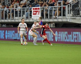 A competitive soccer match is underway with players vying for the ball near the sideline. The players wear different uniforms, indicating they are from opposing teams. The audience is visible in the stadium seating above, and there are visible advertisements and hashtags on the LED boards along the field.