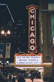 A brightly lit theater sign in an urban area displaying show information for Meghan Trainor and X Ambassadors. The iconic vertical 'Chicago' sign is fully illuminated, contrasting against the night sky. Nearby streetlights and building lights add to the vibrant nighttime cityscape.
