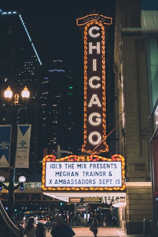 A brightly lit theater sign in an urban area displaying show information for Meghan Trainor and X Ambassadors. The iconic vertical 'Chicago' sign is fully illuminated, contrasting against the night sky. Nearby streetlights and building lights add to the vibrant nighttime cityscape.
