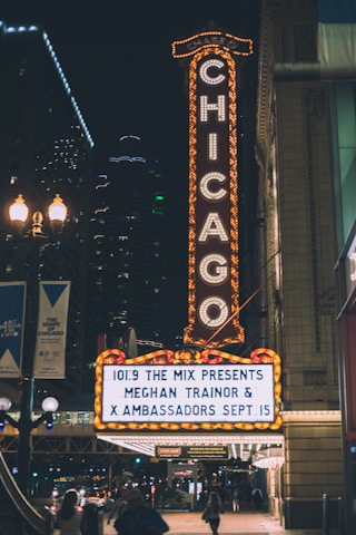 A brightly lit theater sign in an urban area displaying show information for Meghan Trainor and X Ambassadors. The iconic vertical 'Chicago' sign is fully illuminated, contrasting against the night sky. Nearby streetlights and building lights add to the vibrant nighttime cityscape.