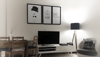 A modern living room with minimalistic decor features a small white entertainment unit supporting a black flat-screen TV. Above the TV, there are three framed posters with simple graphics and motivational text. A wooden dining table with two chairs is placed on the left side, and a cozy armchair with a decorative cushion sits on the right. A tall floor lamp with a black shade provides soft lighting from the corner.