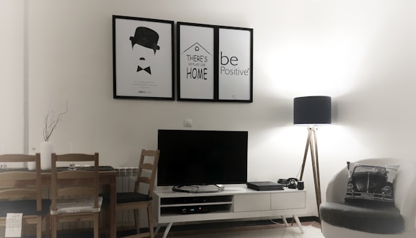 A modern living room with minimalistic decor features a small white entertainment unit supporting a black flat-screen TV. Above the TV, there are three framed posters with simple graphics and motivational text. A wooden dining table with two chairs is placed on the left side, and a cozy armchair with a decorative cushion sits on the right. A tall floor lamp with a black shade provides soft lighting from the corner.