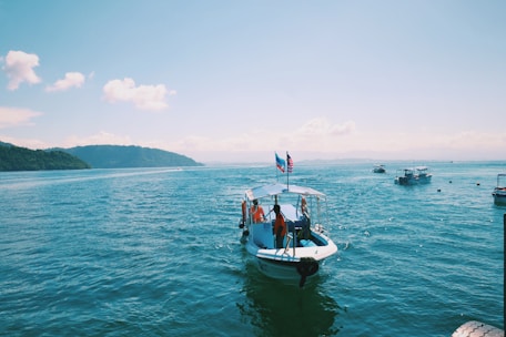 A small boat with a few people on board navigates through bright blue waters, with flags raised at the rear. Several other boats are scattered across the expansive ocean, and distant islands and cloudy skies create a picturesque backdrop.