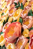 A close-up view of intricately crafted paper flowers with vibrant colors and detailed layers. The flowers have shades of pink, peach, and yellow petals with detailed stamen in the center, highlighted by green leaves.
