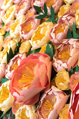 A close-up view of intricately crafted paper flowers with vibrant colors and detailed layers. The flowers have shades of pink, peach, and yellow petals with detailed stamen in the center, highlighted by green leaves.