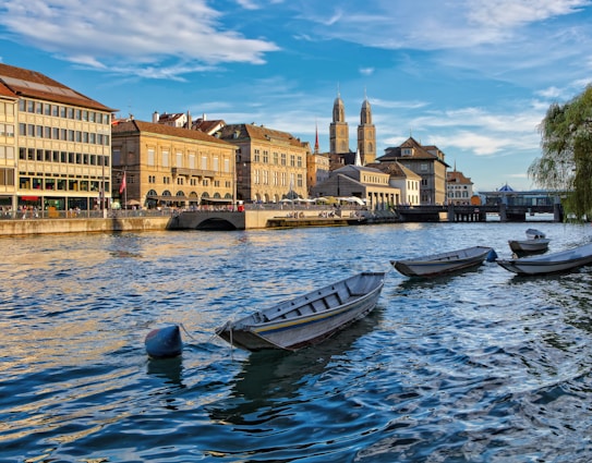 A picturesque waterfront scene features a row of boats gently floating on a river with rippling water. Along the opposite bank, historic buildings with classic architecture line the riverside. Two tall church towers rise prominently in the background, while a bridge crosses the river further down. The setting sun casts warm, golden light on the buildings and water, adding to the serene atmosphere.