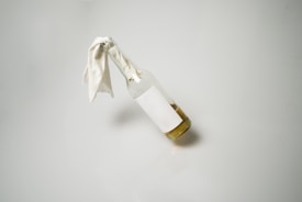 A clear glass bottle positioned horizontally with a white cloth inserted into its mouth. The bottle contains a light brown liquid, and both the bottle and cloth cast a soft shadow on a neutral background.