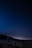 A person stands in a grassy field at night, holding a telescope and gazing up at a starry sky. The horizon is visible with a faint glow, suggesting either a distant village or the remnants of sunset. Trees form a dark silhouette in the background.