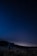 A person stands in a grassy field at night, holding a telescope and gazing up at a starry sky. The horizon is visible with a faint glow, suggesting either a distant village or the remnants of sunset. Trees form a dark silhouette in the background.