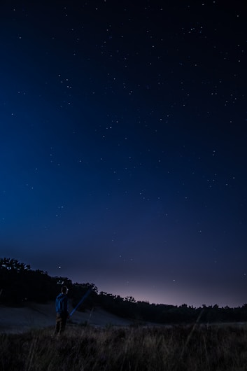A person stands in a grassy field at night, holding a telescope and gazing up at a starry sky. The horizon is visible with a faint glow, suggesting either a distant village or the remnants of sunset. Trees form a dark silhouette in the background.