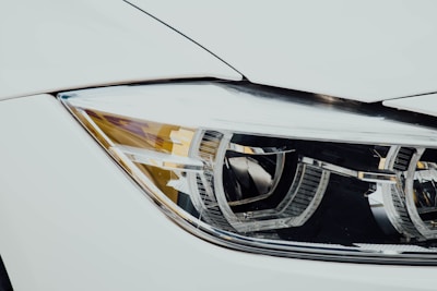 Close-up of a car's headlight featuring a modern, sleek design with intricate reflective elements. The headlight is set against a white car body, highlighting the shiny, metallic components.