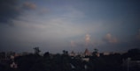 A city skyline with a prominent temple structure in the center, surrounded by lush greenery and urban buildings. The sky is expansive with scattered clouds, casting a serene ambiance over the cityscape.