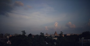 A city skyline with a prominent temple structure in the center, surrounded by lush greenery and urban buildings. The sky is expansive with scattered clouds, casting a serene ambiance over the cityscape.