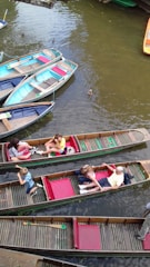 Several wooden boats float on a calm river with a few ducks swimming nearby. People are seated in the boats, enjoying a leisurely boat ride. The boats are painted in various colors including blue, red, and green. The water appears brownish, likely reflecting the natural surroundings.