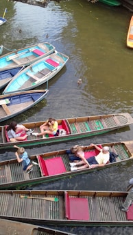 Several wooden boats float on a calm river with a few ducks swimming nearby. People are seated in the boats, enjoying a leisurely boat ride. The boats are painted in various colors including blue, red, and green. The water appears brownish, likely reflecting the natural surroundings.