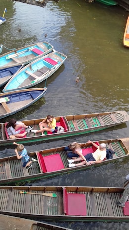 Several wooden boats float on a calm river with a few ducks swimming nearby. People are seated in the boats, enjoying a leisurely boat ride. The boats are painted in various colors including blue, red, and green. The water appears brownish, likely reflecting the natural surroundings.