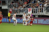 A women's soccer match is taking place with players in action near the sideline. Three players in white jerseys are engaged with a player in a red and black jersey who is sliding on the grass. A referee in a yellow shirt watches nearby, and a crowd is visible in the stands. The stadium is equipped with banners and digital signage displaying brand names and social media hashtags.