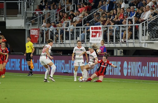 A women's soccer match is taking place with players in action near the sideline. Three players in white jerseys are engaged with a player in a red and black jersey who is sliding on the grass. A referee in a yellow shirt watches nearby, and a crowd is visible in the stands. The stadium is equipped with banners and digital signage displaying brand names and social media hashtags.