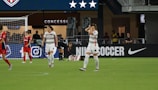 A soccer match scene shows players on a field, some in red and others in white uniforms. One player is injured and lying on the ground while another kneels beside them. The area behind features a concession stand with a large advertisement for Nike Soccer. Spectators and staff are visible in the background.