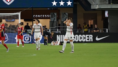 A soccer match scene shows players on a field, some in red and others in white uniforms. One player is injured and lying on the ground while another kneels beside them. The area behind features a concession stand with a large advertisement for Nike Soccer. Spectators and staff are visible in the background.